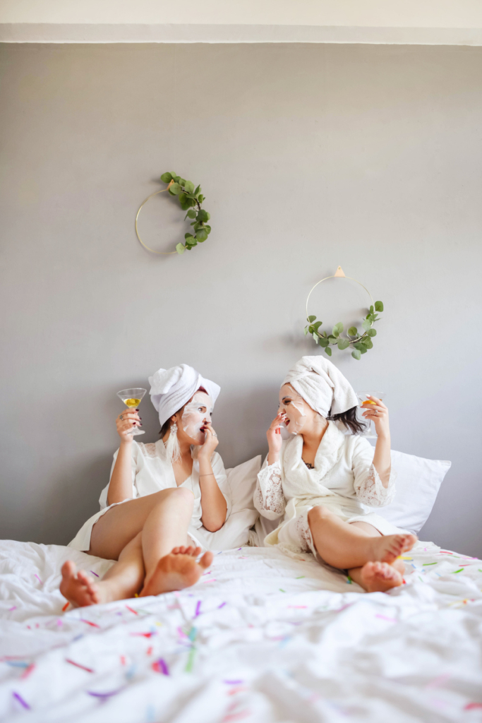 Barátnők arcmaszkban, koktéllal a kezükben ülnek az ágyon