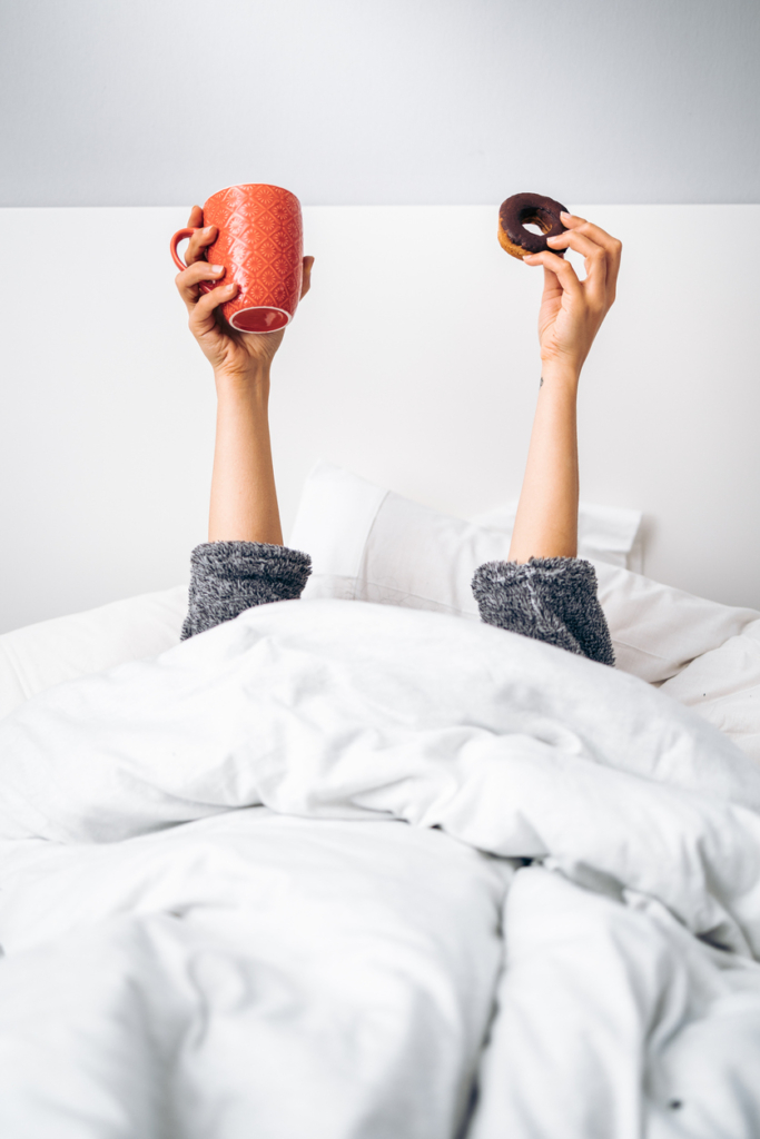 Woman lying in bed holding a coffee mug in one hand and a vanilla ring cookie in the other