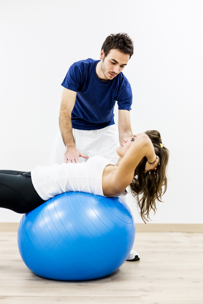 Nő egy nagy gumilabdán gyógytornász segítségével tornázik