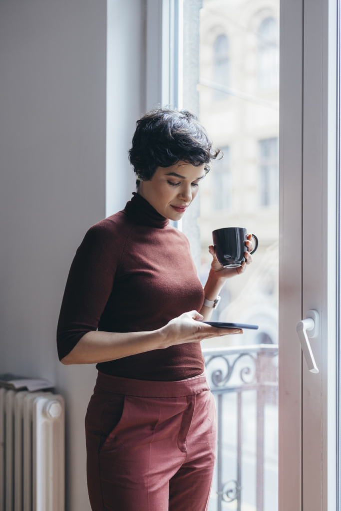 Nő az ablaknál nyomkodja a telefonját, másik kezében egy bögre tea