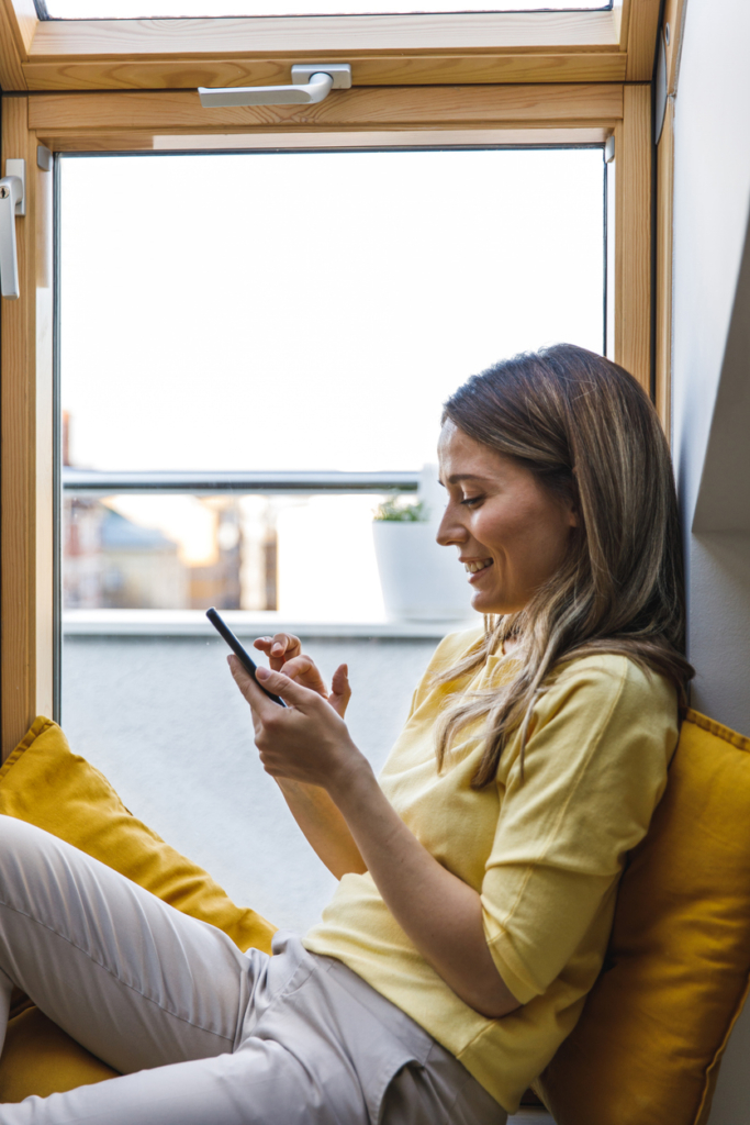 Nő az ablakban ülve olvasgat a mobiltelefonján