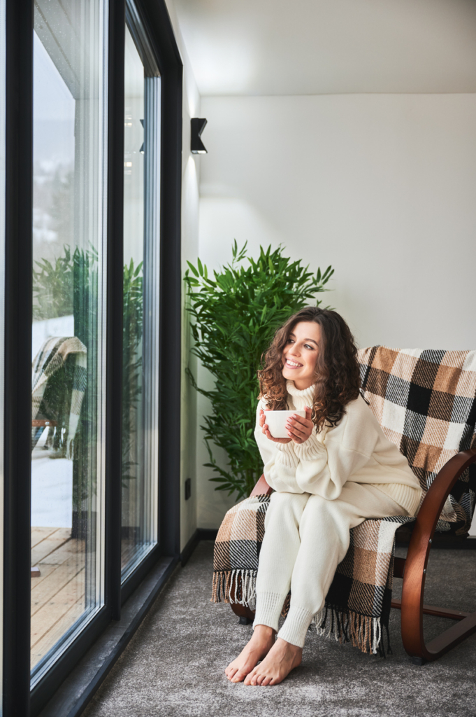 Fiatal nő otthon, meleg takaróval fedett fotelből néz ki a hatalmas üvegablakokon