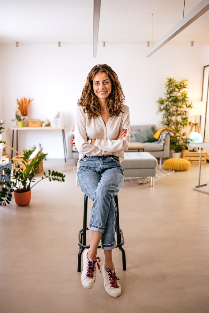 Portré egy nőről, aki egy kisszéken ül, keresztbe font karokkal és lábakkal