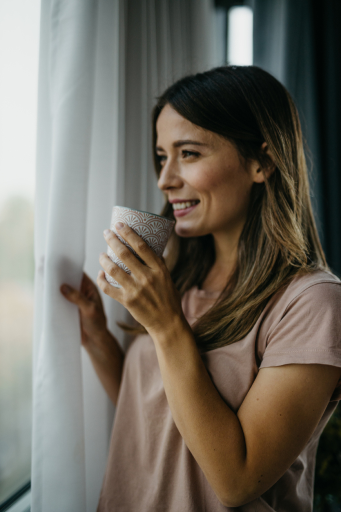 Nő mosolyogva iszik egy kávét az ablaknál