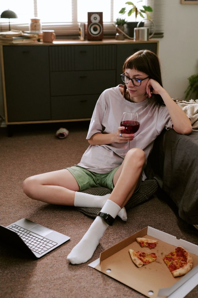 Nő borral és pizzával a kezében ül a nappalija padlóján a laptop előtt