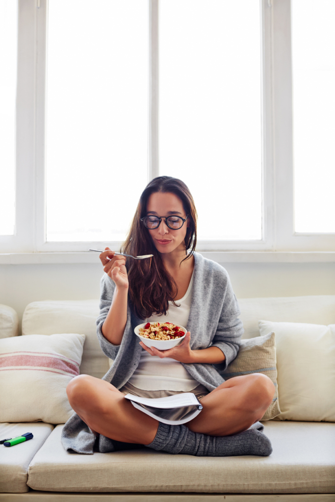 Egészséges reggelijét fogyasztja egy szemüveges fiatal nő a kanapén ülve