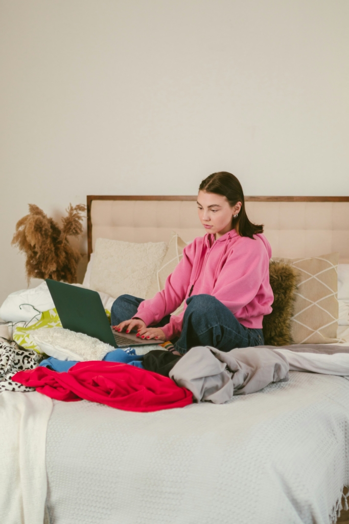 Nő laptoppal ül az ágyon, körülötte ruhák hevernek szerte szét