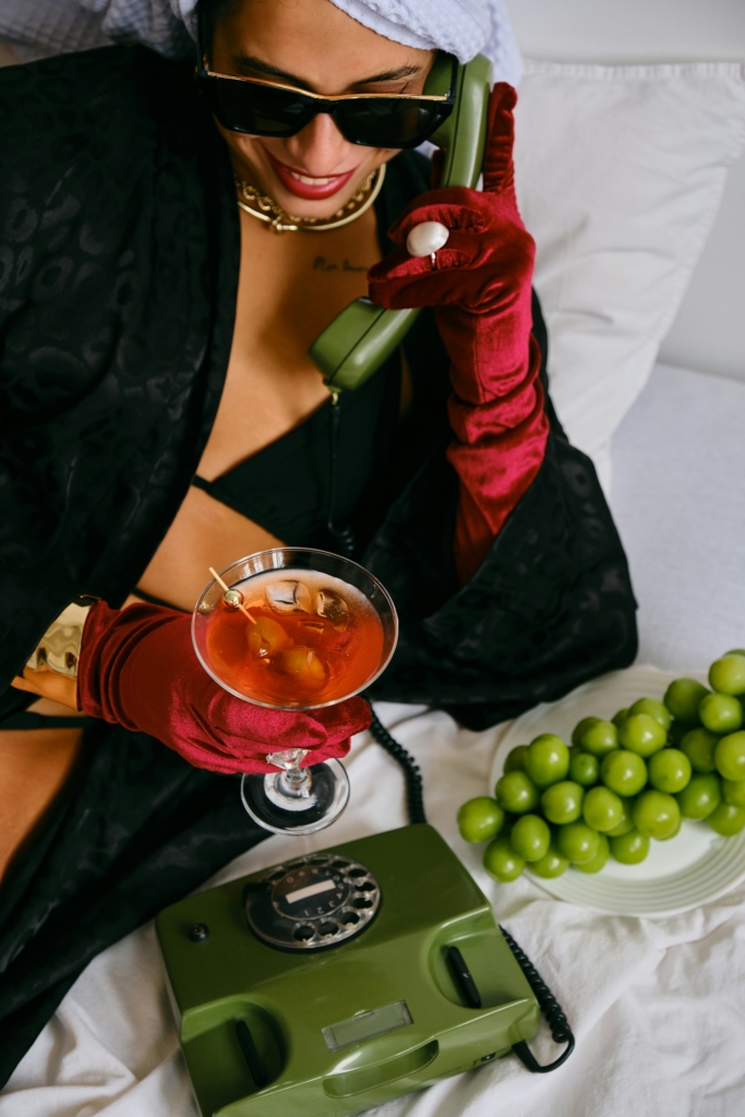 Nő bőrkesztyűben telefonál és koktélozik egy szállodai ágyban