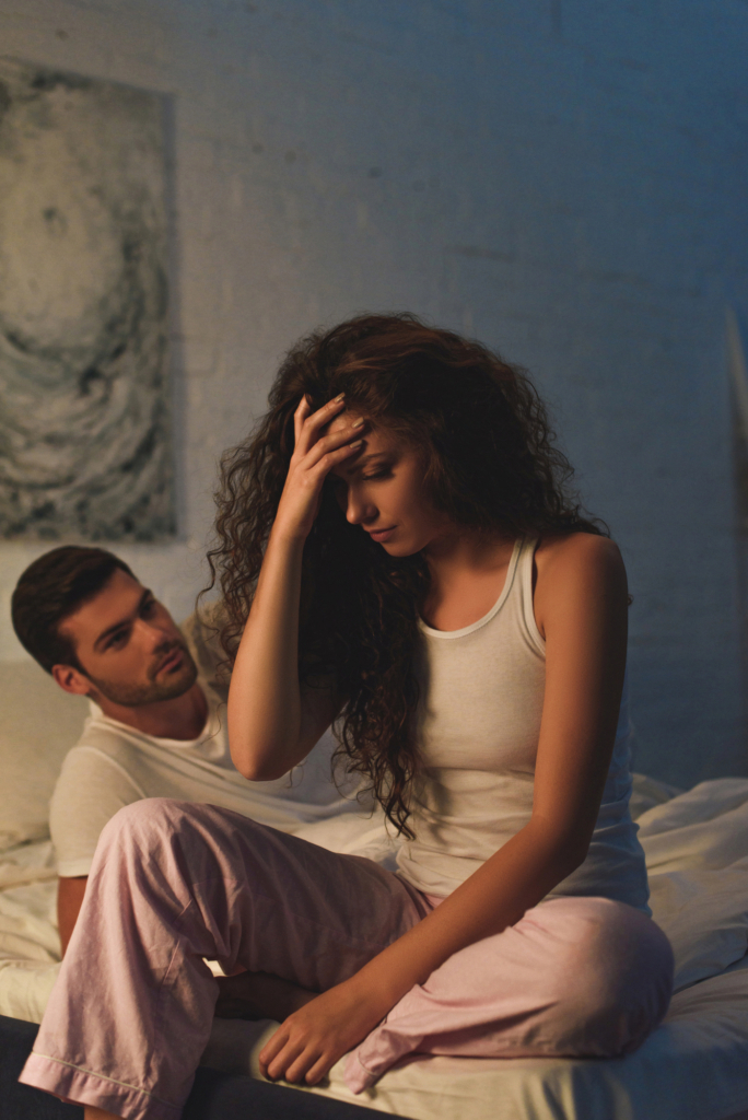 Nő az ágyon ül és fogja a fejét, a párja kérdőn néz rá