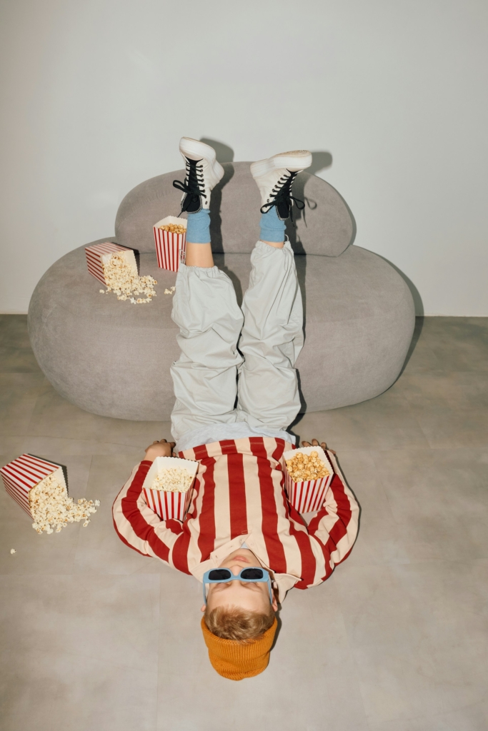Kisfiú fejjel lefelé fekszik egy fotelben, körülötte csomó popcorn