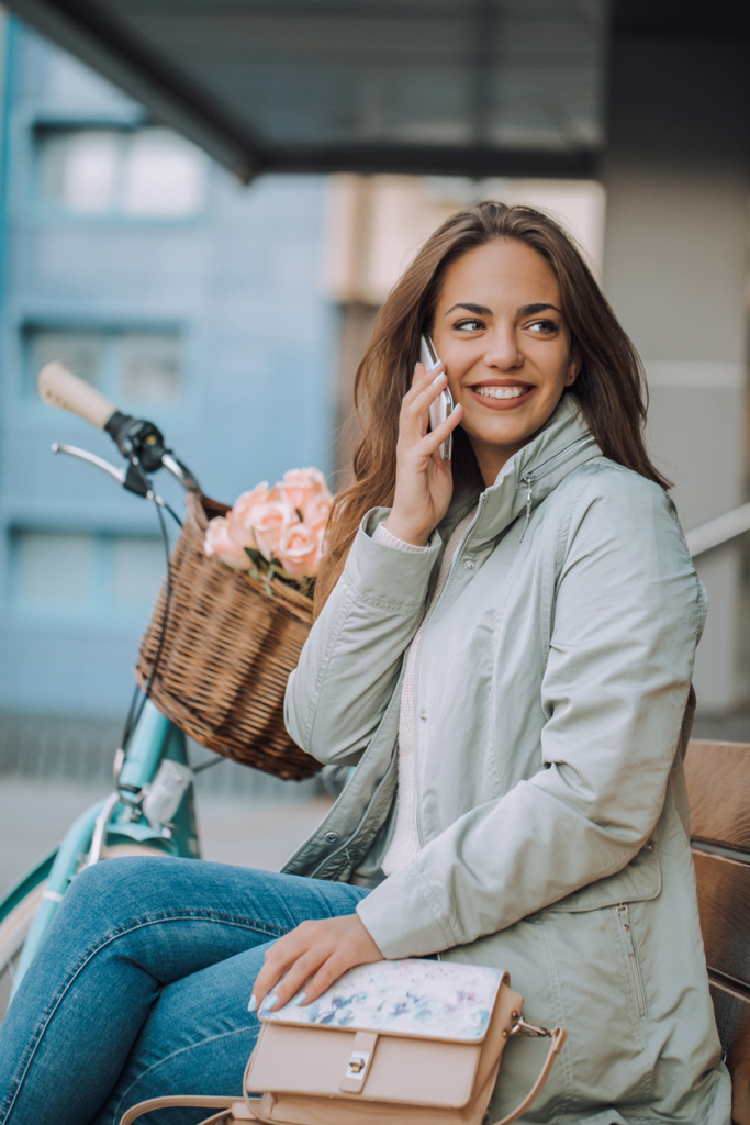 Mosolygós fiatal nő mobiltelefonál egy padon ülve, mellette a biciklije letámasztva