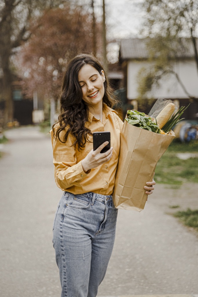 Nő bevásárlásból sétál haza, kezében a bevásárlás és a mobiltelefonja