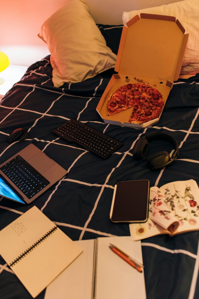Pizza, laptop, füzetek, könyvek, tablet és fejhallgató az ágyra dobva