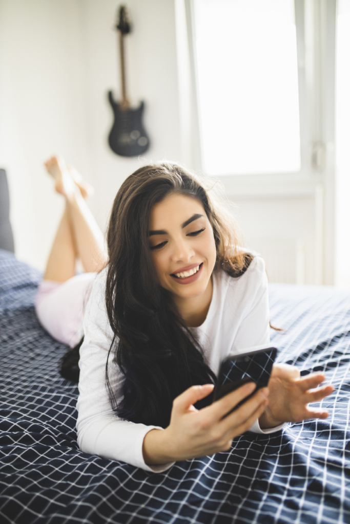 Joven sonriente mirando el teléfono tumbada en la cama