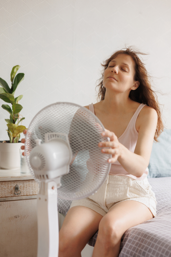 Nő az ágy szélén ül és fogja az álló ventilátor szélét