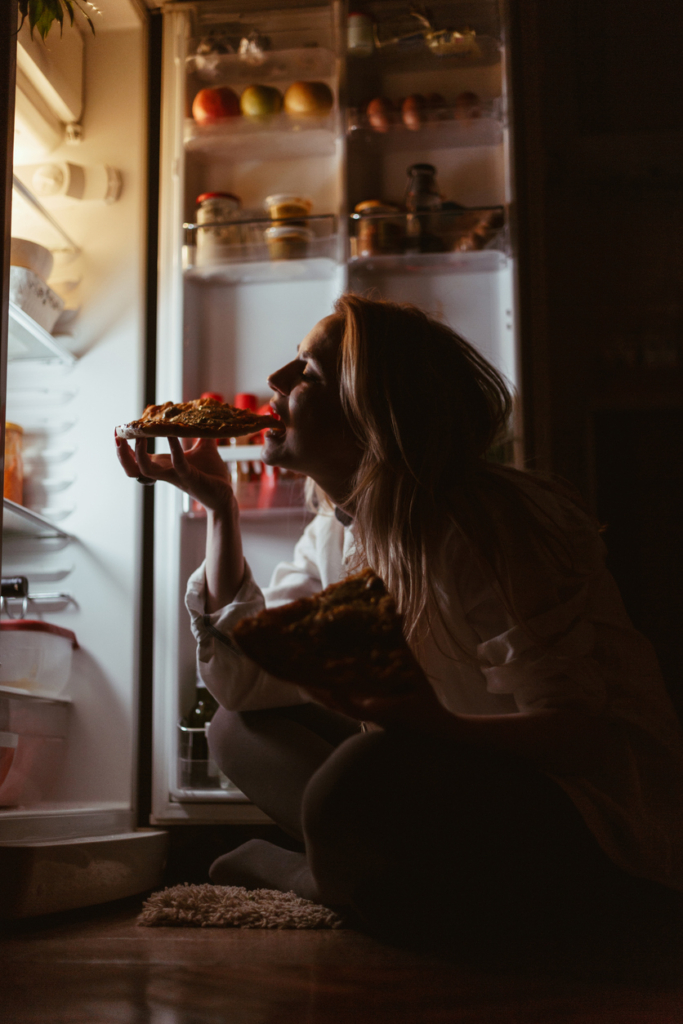 Nő a hűtőszekrény előtt ülve a sötétben megeszik egy szelet pizzát