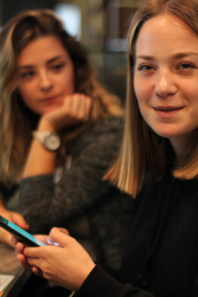 Két barátnő egy kávézóban, az egyik a telefonját tartja a kezében, a másik csendben ül